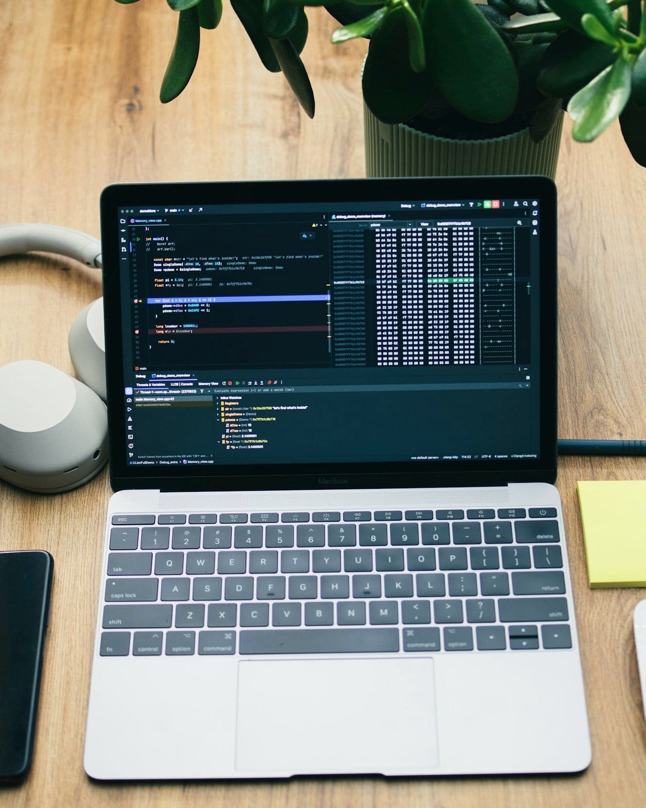 Computadora portátil con código en pantalla sobre un escritorio de madera, junto a auriculares y una planta.